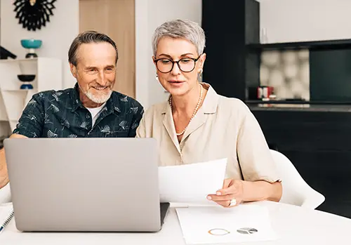 Senior retired couple at a table in dining room using a laptop computer. Two mature people looking at paperwork and discussing their Medicare options